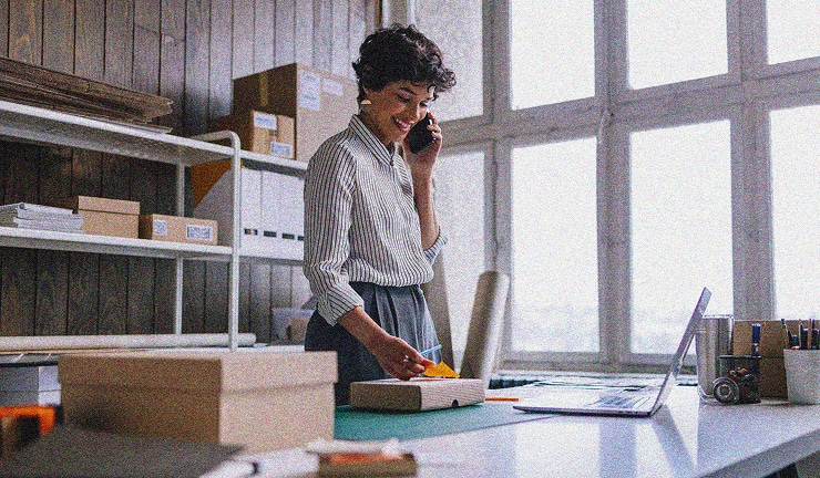 A woman stands at a desk in a bright office, talking on the phone and writing in a notebook. Boxes, shelves, and a laptop are visible around her, suggesting she is working in a small business or studio.
